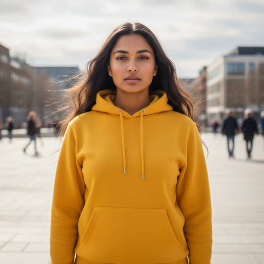 Female model wearing a golden yellow hoodie standing outdoors with blurred people and buildings in the background. Plain Golden Yellow Hoodie by Paryatak Travel Shop.