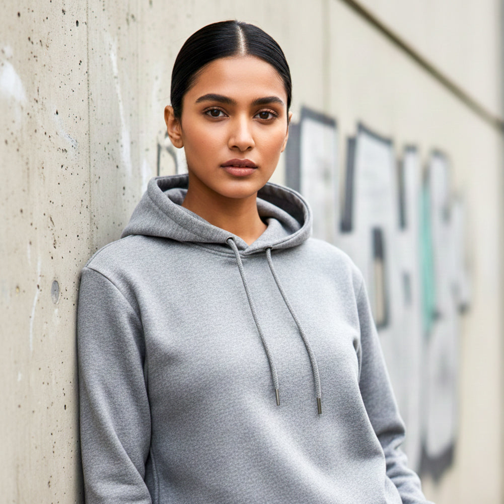 Woman model wearing a melange grey hoodie leaning against a concrete wall. Plain Melange Grey Hoodie by Paryatak Travel Shop.